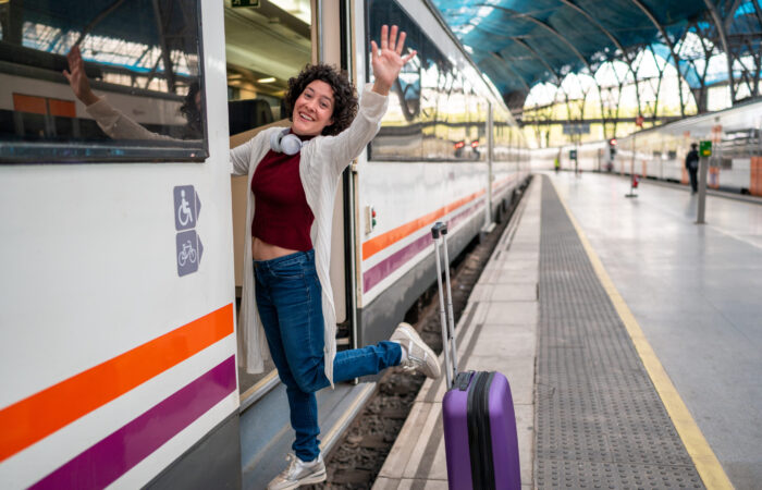 Young woman waving goodbye while getting off a train at a railway station with her trolley